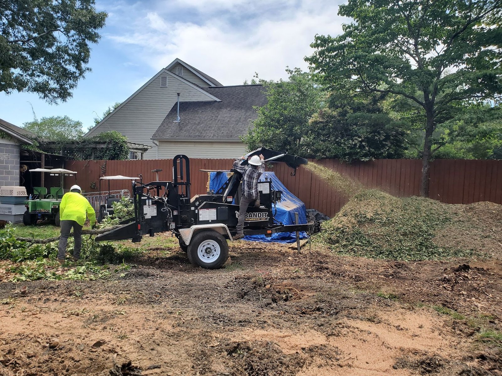 Crew member atop the chipper trailer feeding brush while a coworker rakes debris near a privacy fence